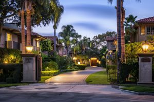A serene residential street at sunset with palm trees and manicured lawns. The brick-paved street is lined with well-lit, elegant homes. Warm outdoor lighting enhances the peaceful ambiance, while an armed security guard from San Francisco County stands by a wrought iron gate that invites passersby in.