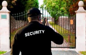 A security guard, seen from behind, speaks into a handheld radio while standing in front of a large, closed iron gate framed by stone pillars. Trees and a pathway are visible behind the gate. The word "SECURITY" is written in white on the back of the guard's black jacket. This scene depicts residential security in San Francisco County.