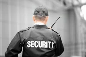 A person wearing a black security uniform and cap is seen from behind, holding a walkie-talkie. The background is slightly blurred, highlighting the individual as they patrol or stand guard near a building. This scene could easily depict someone involved in Residential Security San Francisco County.